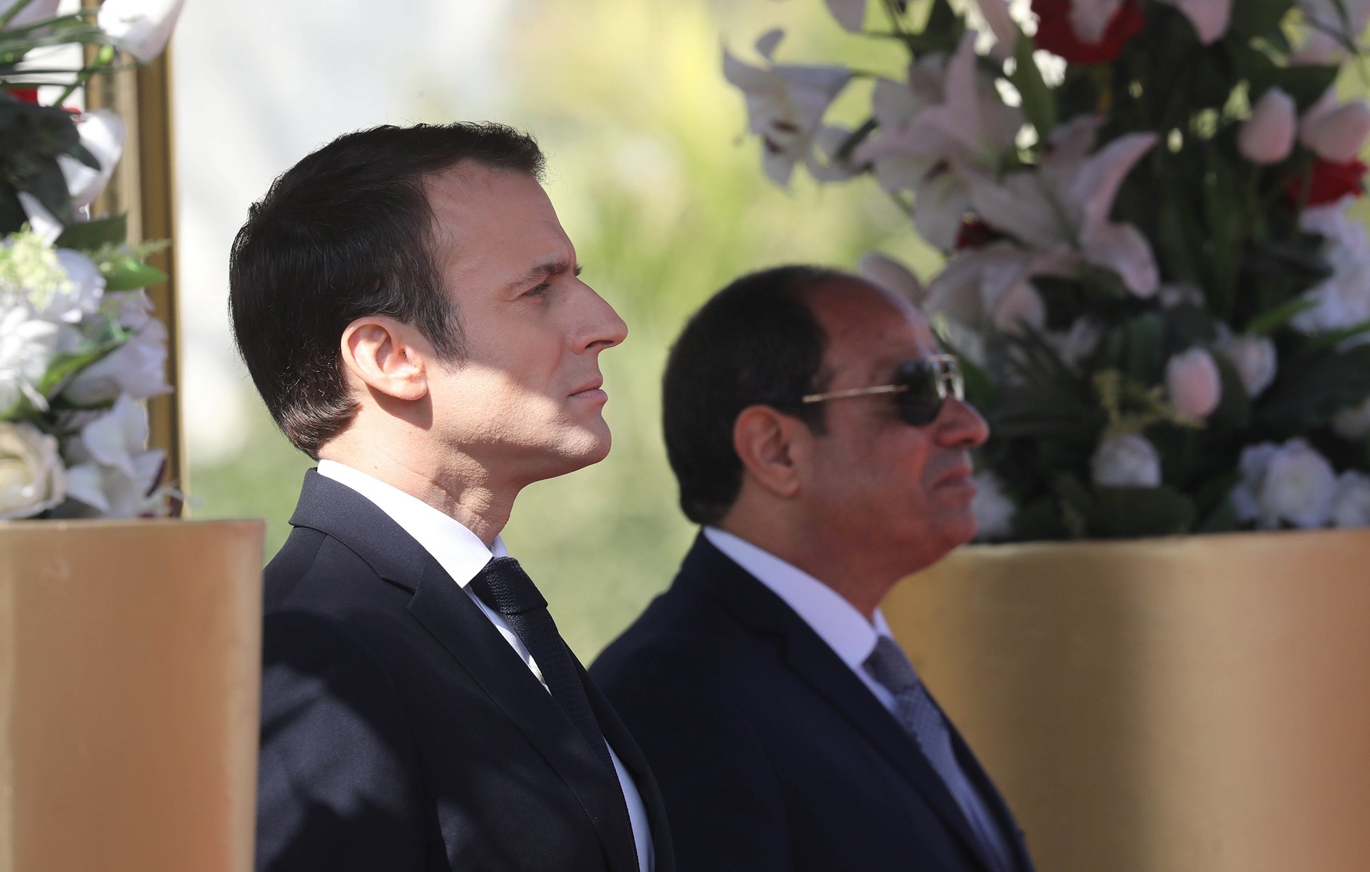 الصورة تظهر رجلين standing بجانب بعضهما البعض. الرجل على اليسار يرتدي بدلة داكنة مع ربطة عنق، بينما الرجل على اليمين يرتدي نظارات شمسية وبدلة رسمية. الخلفية تحتوي على زهور وأشجار، مما يضفي جوًا من الاحتفال أو المناسبة الرسمية. تعكس الصورة موقفًا رسميًا وجديًا بين الشخصين.