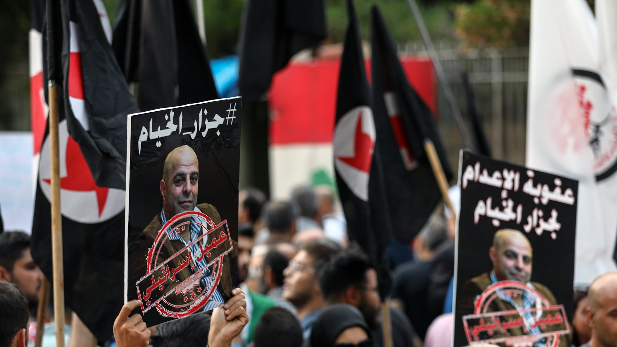 الصورة توضح تجمعًا حاشدًا حيث يحمل المتظاهرون لافتات تحمل عبارات وشعارات مختلفة. من بين اللافتات، توجد صور لشخص يبدو أنه شخصية بارزة، مع وجود علامة تشير إلى معارضة أو احتجاج يتعلق بموضوع الإعدام. المتظاهرون يرتدون ملابس متنوعة، وبعضهم يحمل أعلامًا. الأجواء تعكس شعوراً بالاحتجاج والرفض.