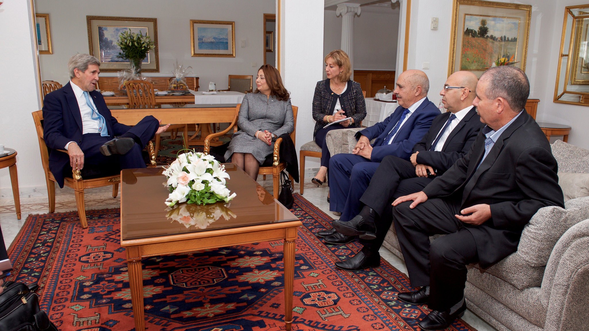 تظهر الصورة مجموعة من الأشخاص يجلسون في غرفة معيشة أنيقة. في المنتصف، يوجد طاولة قهوة تحمل زهورًا بيضاء. يبدو أن هناك نقاشًا أو اجتماعًا يجري بينهم. يجلس شخص يرتدي بدلة زرقاء في الجهة اليسرى، بينما يجلس الآخرون على الأرائك المحيطة. الخلفية تحتوي على لوحات فنية ونباتات، مما يضفي جوًا مريحًا على المكان.