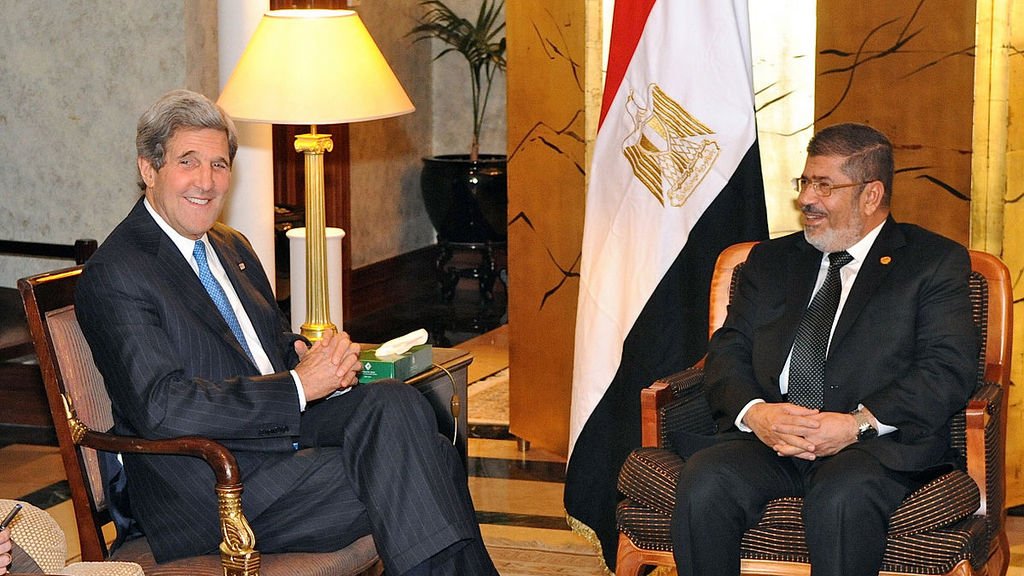 تظهر الصورة اجتماعًا بين شخصين يجلسان في غرفة مريحة. يجلس أحدهما على اليسار وهو يرتدي بدلة رسمية مع ربطة عنق ويبتسم، بينما يجلس الشخص الآخر على اليمين، كذلك يرتدي بدلة ويبدو سعيدًا. الخلفية تحتوي على علم مصر، مما يشير إلى أن الاجتماع قد يكون ذو طابع رسمي يتعلق بالشؤون السياسية أو العلاقات الدولية. الأجواء تبدو ودية وإيجابية.