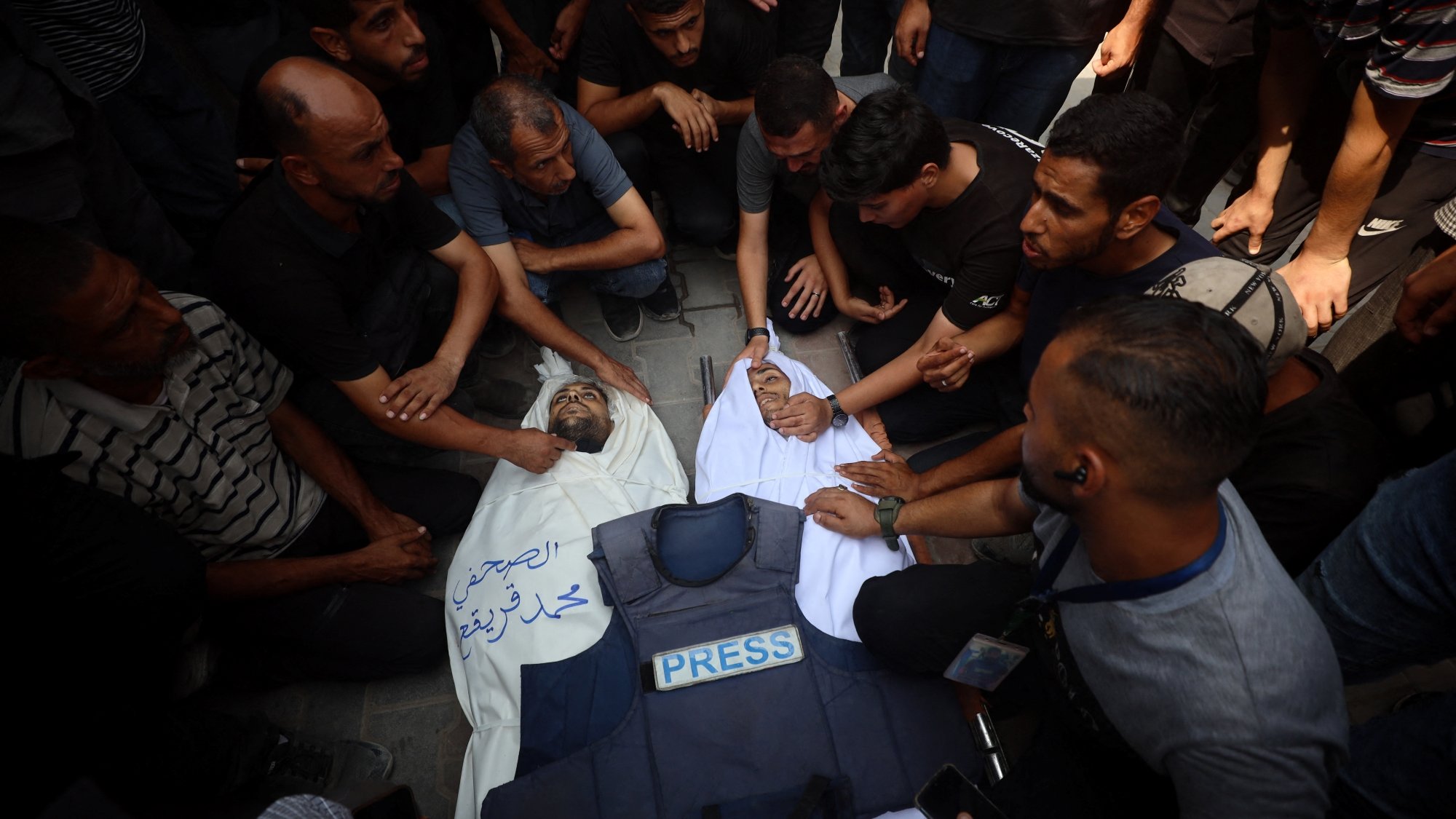 A group of people mourns over a deceased individual in a white shroud, surrounded by a press vest.