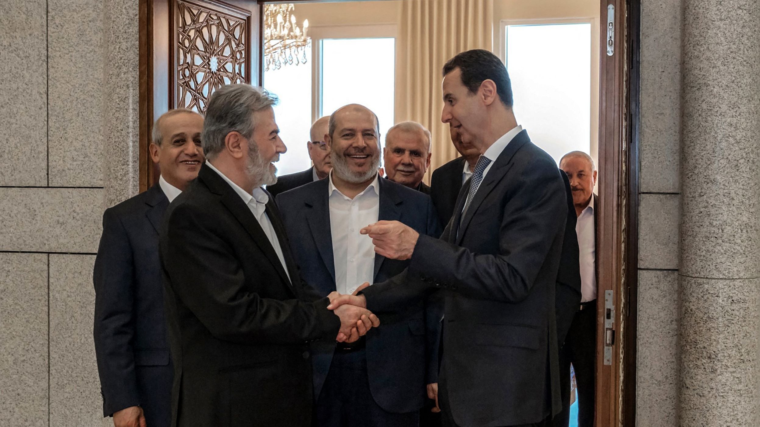 تظهر الصورة مجموعة من الرجال في قاعة استقبال. يتوسط الصورة شخصان يتصافحان، حيث يبدو أن أحدهما في وضعية حديث مع الآخر. الخلفية تحتوي على أبواب وزخرفة رائعة، وهناك أشخاص آخرون يقفون خلفهم، يظهر عليهم تفاعل إيجابي ورغبة في التواصل. الأجواء تبدو رسمية، مما يشير إلى لقاء مهم أو مناسبة خاصة.