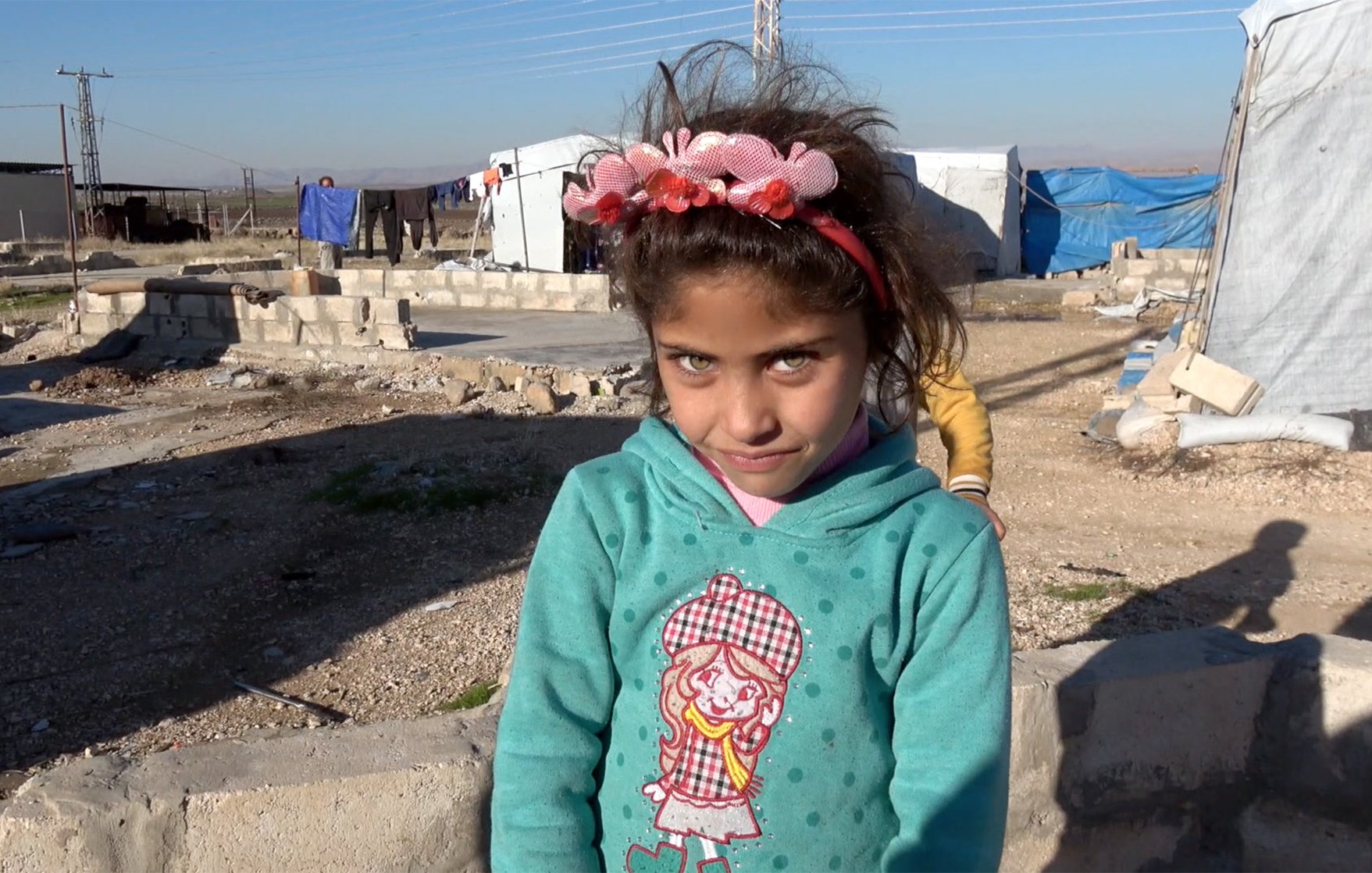 تظهر في الصورة فتاة صغيرة ترتدي سترة زرقاء مزينة بصورة لفتاة أخرى. لديها عيون فاتحة ومشرقة وشعر داكن، مع زينة زهرية في شعرها. الخلفية تضم خيام وغير منظمة، مما يوحي بأنها في مخيم. تبدو الفتاة مبتسمة ومرحة.