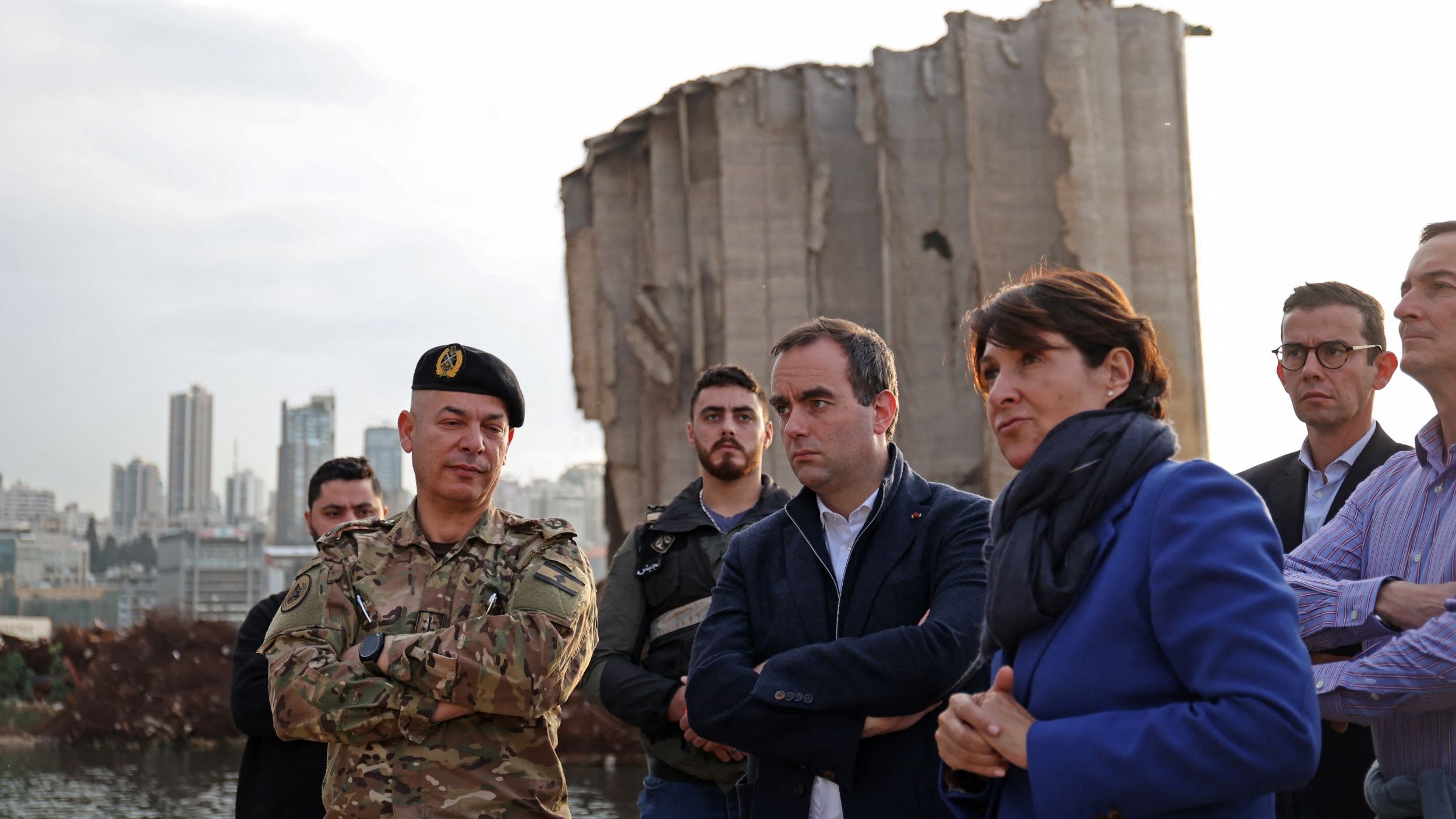 تظهر في الصورة مجموعة من الأشخاص في منطقة تضررت من انفجار أو حدث كارثي. في المقدمة، يوجد ضابط عسكري يرتدي زيًا رسميًا ولديه شارة على القبعة. بجانبه، يقف مجموعة من الرجال والنساء يتحدثون بجدية، مع تعابير وجه تعكس الانتباه والتفكير. في الخلفية، توجد آثار دمار واضحة، مع مبنى شاهق متضرر بشكل كبير. السماء تميل إلى اللون الرمادي، مما يضفي أجواءً من الحزن أو التأمل على المشهد.