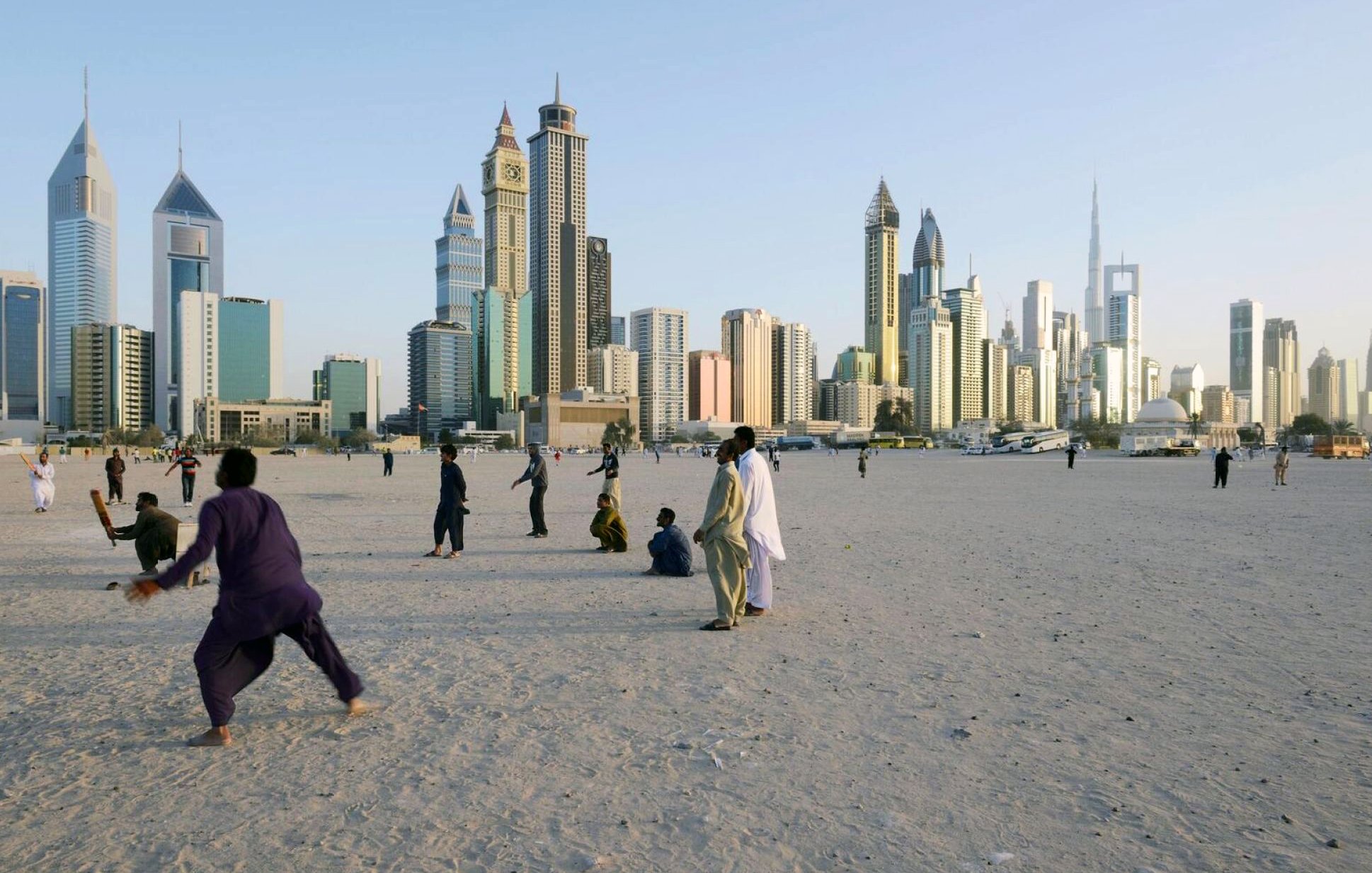 الصورة تُظهر مشهدًا حضريًا لمدينة حديثة، حيث تبرز ناطحات السحاب والمباني الشاهقة. في المقدمة، يوجد مجموعة من الأشخاص يمارسون أنشطة مختلفة على رمال منطقة مفتوحة، ما يوحي بأجواء حيوية. الخلفية تُظهر المدينة مع سماء زرقاء تتخللها أشعة الشمس، مما يضفي لمسة جميلة على المشهد.