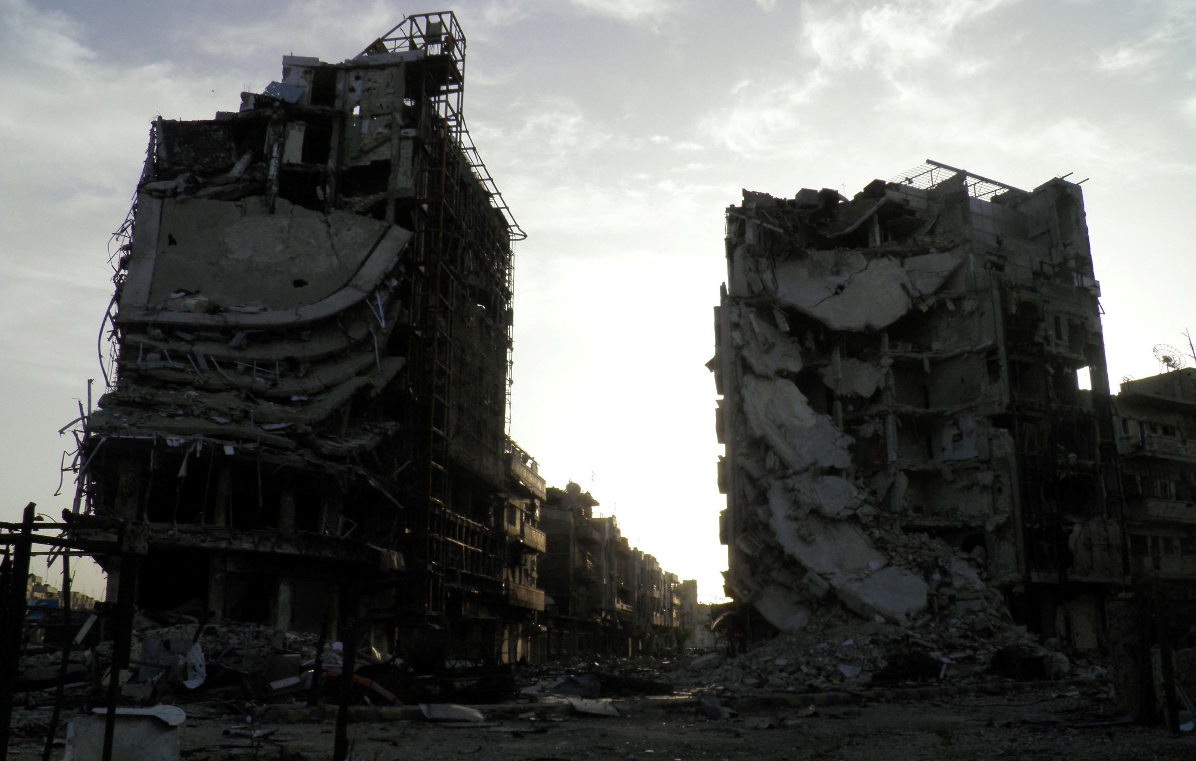 تظهر الصورة عمارتين محطمتين في منطقة يبدو أنها تعرضت لتدمير واسع. المباني مشوهة، حيث تظهر الجدران المنهارة والأسطح المدمرة، مما يعكس آثار الدمار. السماء في الخلفية تبدو ملبدة، مما يضيف شعورًا بالخيبة والخراب. المنطقة المحيطة يبدو أنها مهجورة، مما يعكس الأثر العميق للصراعات أو الكوارث التي أدت إلى هذا الوضع.