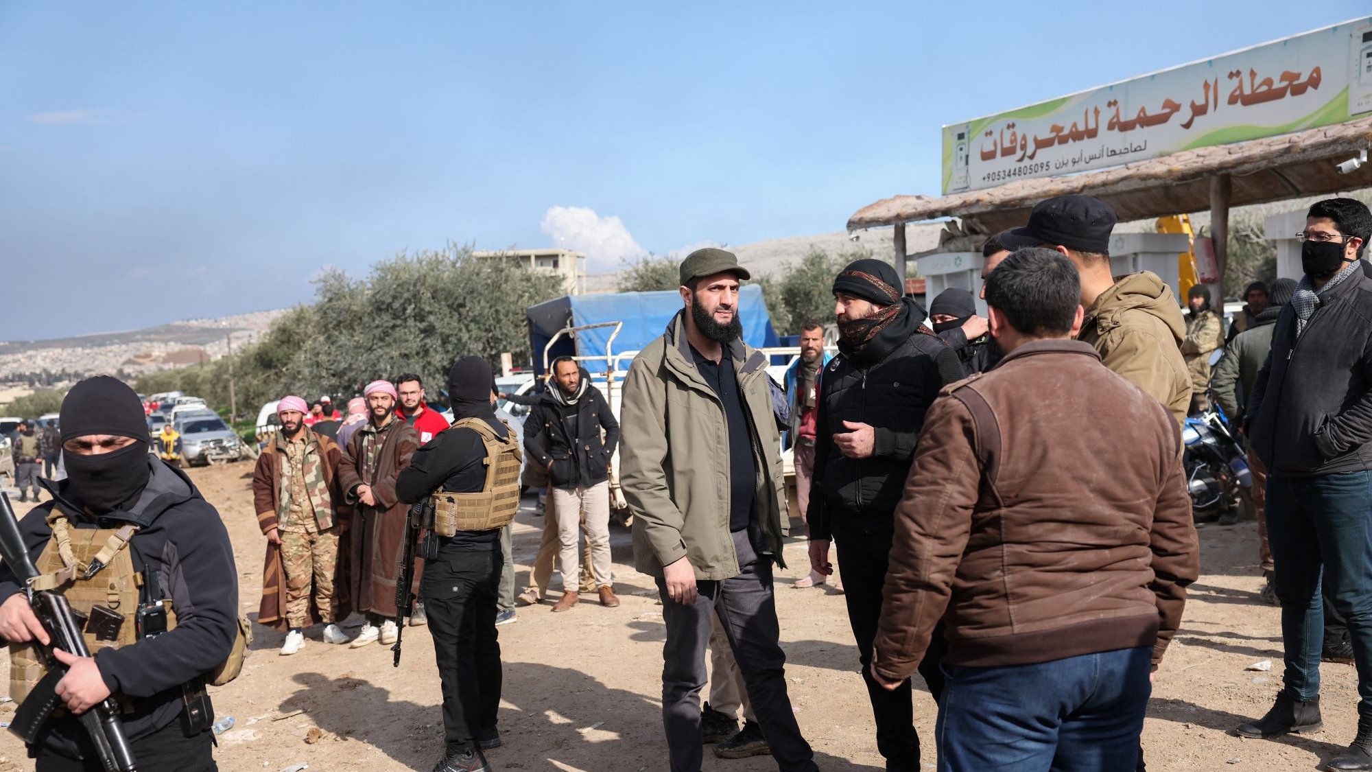 تظهر الصورة مجموعة من الأشخاص في منطقة مفتوحة تحت سماء زرقاء مع بعض الغيوم. يبدو أن هناك عددًا من الرجال يرتدون ملابس تقليدية وبعضهم يرتدي ملابس عسكرية أو شبه عسكرية. في الخلفية، يوجد لافتة تشير إلى "محطة الرحمة للمشاريع". يُلاحظ أن بعض الأشخاص يتحدثون مع بعضهم البعض، وهناك أيضًا مجموعة من الأشخاص يحملون أسلحة. الأجواء تبدو نشطة وتعبر عن تفاعل اجتماعي بين الحضور.