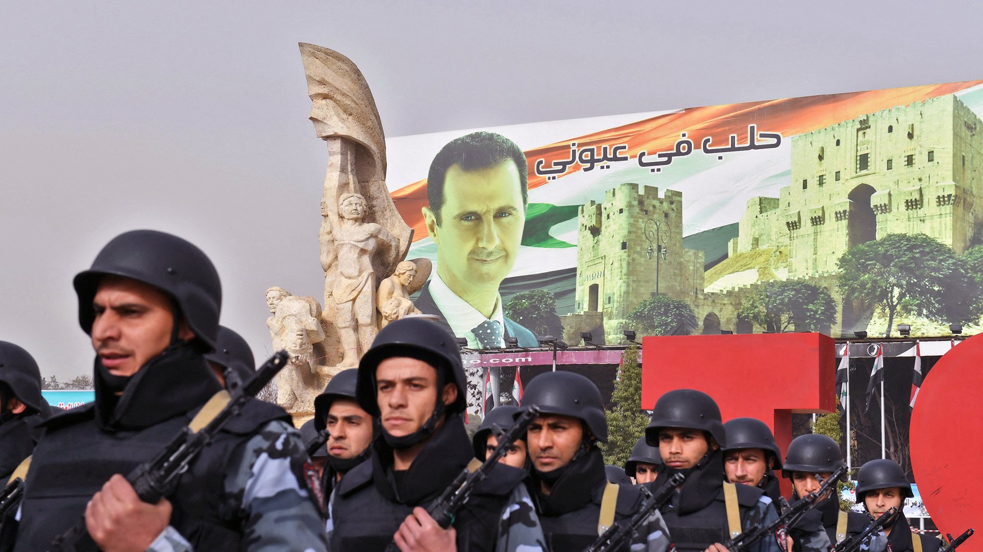 تظهر الصورة عرضًا عسكريًا أو تظاهرة، حيث يسير مجموعة من الجنود في صفوف منظمة، يرتدون زيًا عسكريًا ويحمون أسلحة. خلفهم يوجد لافتة ضخمة تحمل صورة لرجل، تترافق مع مشاهد معمارية تتعلق بسوريا. تظهر أيضًا بعض الرموز الوطنية مثل الأعلام. الأجواء تعكس نوعًا من القوة والتأييد السياسي.