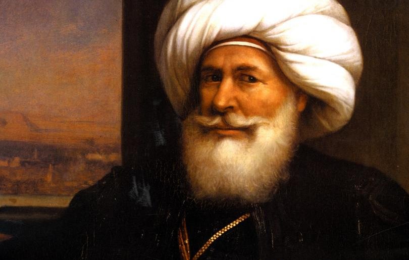 تظهر الصورة رجلاً مسنًّا يرتدي عمامة كبيرة ومجموعة من الملابس التقليدية. يتميز بلحيته البيضاء الكثيفة وابتسامة ودودة. يحمل سيفًا في يده، مما يوحي بمكانته العالية أو دوره القيادي. الخلفية تحتوي على مشهد يبدو كأنه منظر طبيعي لمدينة أو تجمع حضري. تعكس تفاصيل ملابسه وزينه التراث الثقافي لمنطقته.