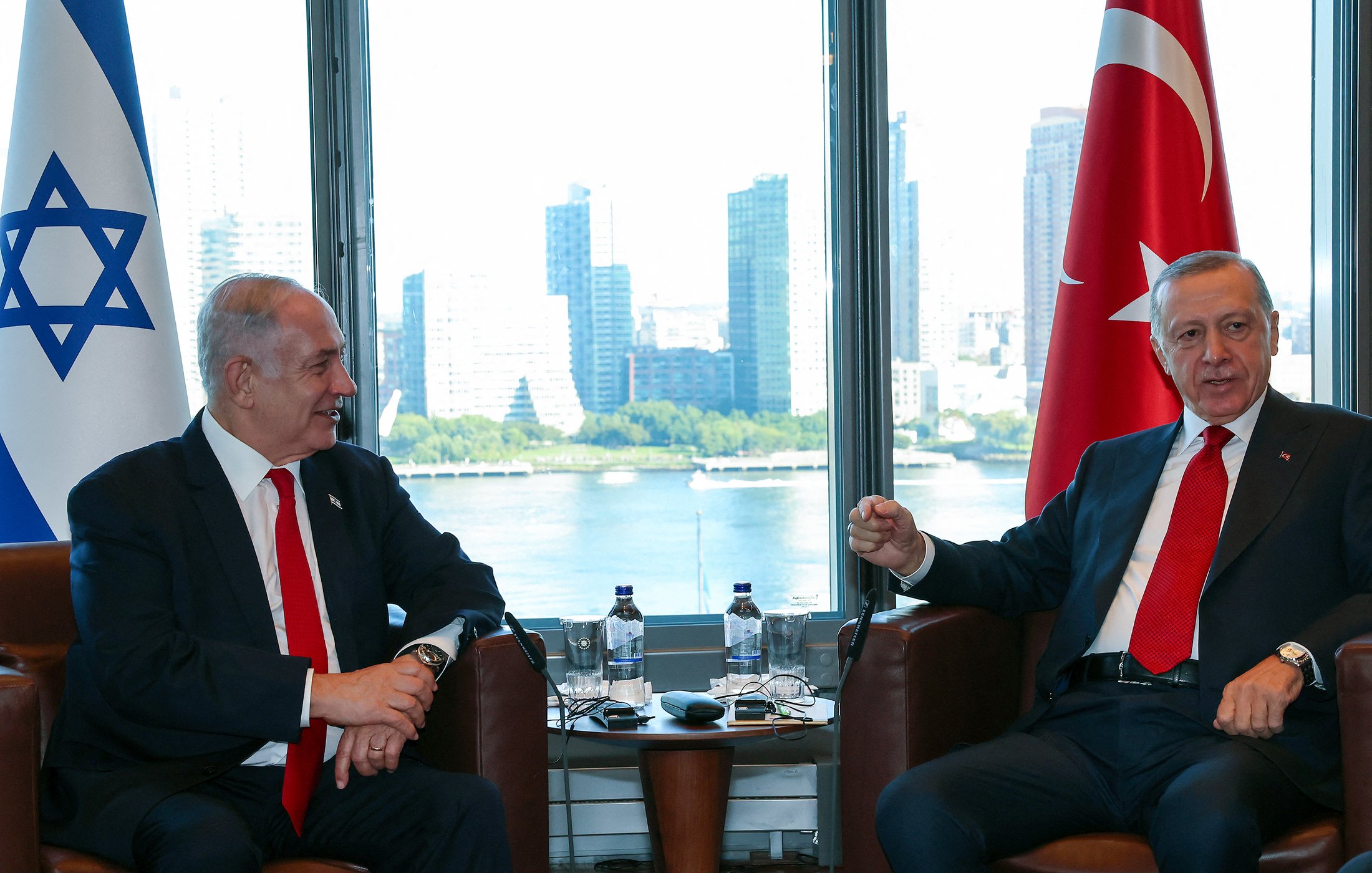 تظهر في الصورة اجتماع بين شخصين يجلسان في غرفة ذات إطلالة على مدينة حديثة. أحدهما يرتدي بدلة داكنة مع ربطة عنق حمراء، والآخر يرتدي بدلة رمادية. خلفهما يوجد علم إسرائيل إلى اليسار وعلم تركيا إلى اليمين. يظهر في الخلفية نهر وبعض المباني الشاهقة، مما يعكس أجواء حضرية حديثة. يبدو أن النقاش يجري في أجواء دبلوماسية.
