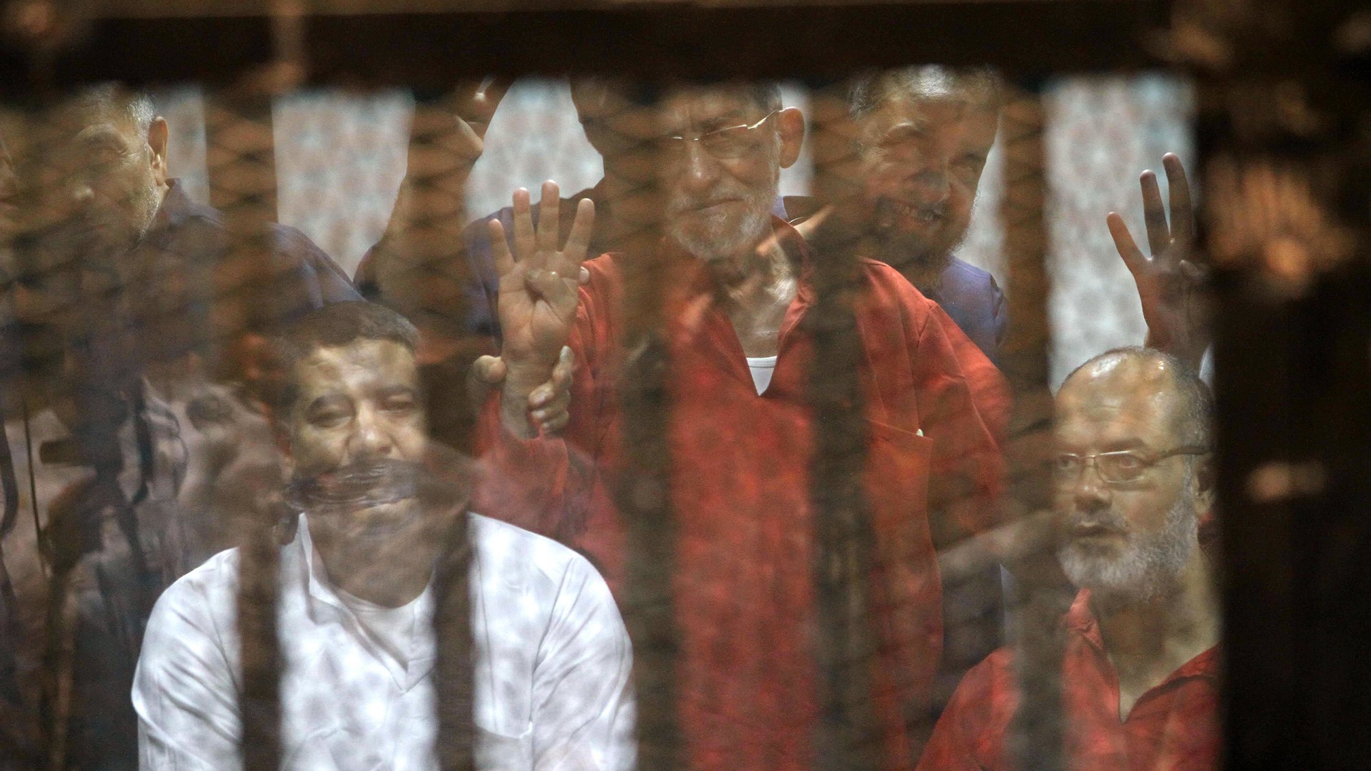 تظهر في الصورة مجموعة من الأشخاص الموجودين خلف قضبان. يبدو أن بعضهم يرفعون إشارة معينة، بينما تعكس الشبكة المحيطة بهم بعض التفاصيل، مما يعطي انطباعًا عن مجموعة محكومة في بيئة موصولة. الألوان تتراوح بين الأحمر والأبيض، مما يشير إلى ملابس يمكن أن تكون متعلقة بسياق خاص. الوضع العام يوحي بالتوتر أو الأحداث الجادة.