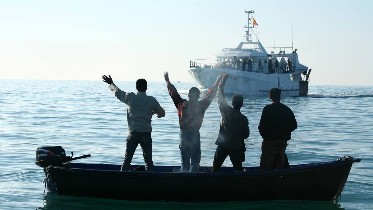 تظهر في الصورة مجموعة من الأشخاص يقفون في قارب صغير في المياه الهادئة، ويبدو أنهم يلوحون بأيديهم نحو قارب أكبر يقترب منهم. المشهد يعكس نشاطًا بحريًا، حيث الأجواء هادئة وتظهر السماء والماء بلونٍ صافٍ. الأشخاص يظهرون من الخلف، مما يركز الانتباه على الإيماءات التي يقومون بها.