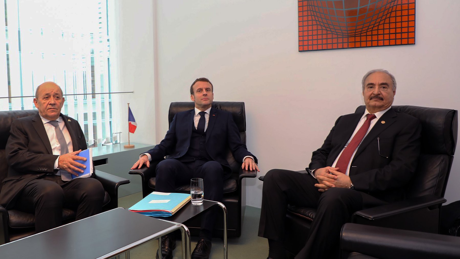 تظهر الصورة ثلاثة رجال يجلسون في غرفة اجتماعات. الشخص في المنتصف يرتدي بدلة داكنة، بينما الأخران يجلسان على جانبيه. يوجد علم فرنسي صغير في الجهة اليسرى من الصورة، والطاولة أمامهم تحتوي على أوراق وزجاج ماء. الجدران محاطة بالزجاج وتحتوي على عمل فني ملون في الأعلى. تعكس التعبيرات والوضوح في الجلسة وجود نقاش أو اجتماعات رسمية.