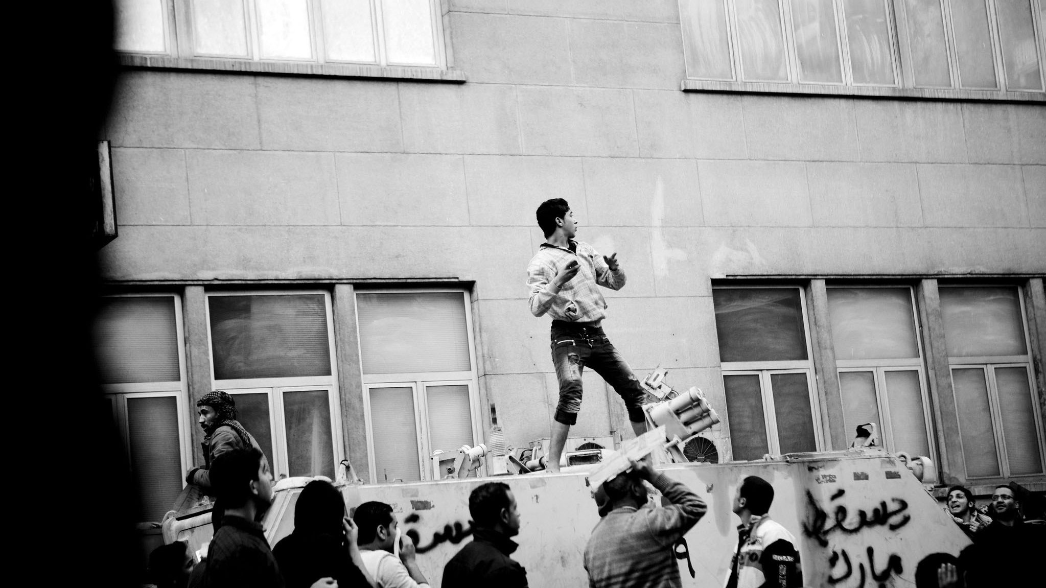 تظهر الصورة مشهداً من تظاهرة أو حدث اجتماعي. في المركز، يوجد شاب يقف على قمة السيارة أو المنصة، عازفاً على آلة موسيقية. يحيط به جمع من الناس، الذين يبدو أنهم يعبّرون عن حماسهم أو تأييدهم. الخلفية تتكون من مبنى بملامح عصرية، والزخارف على السيارة تحمل بعض الكتابات باللغة العربية. الصورة بالأبيض والأسود، مما يضيف عمقاً وأجواء مأساوية للمشهد.