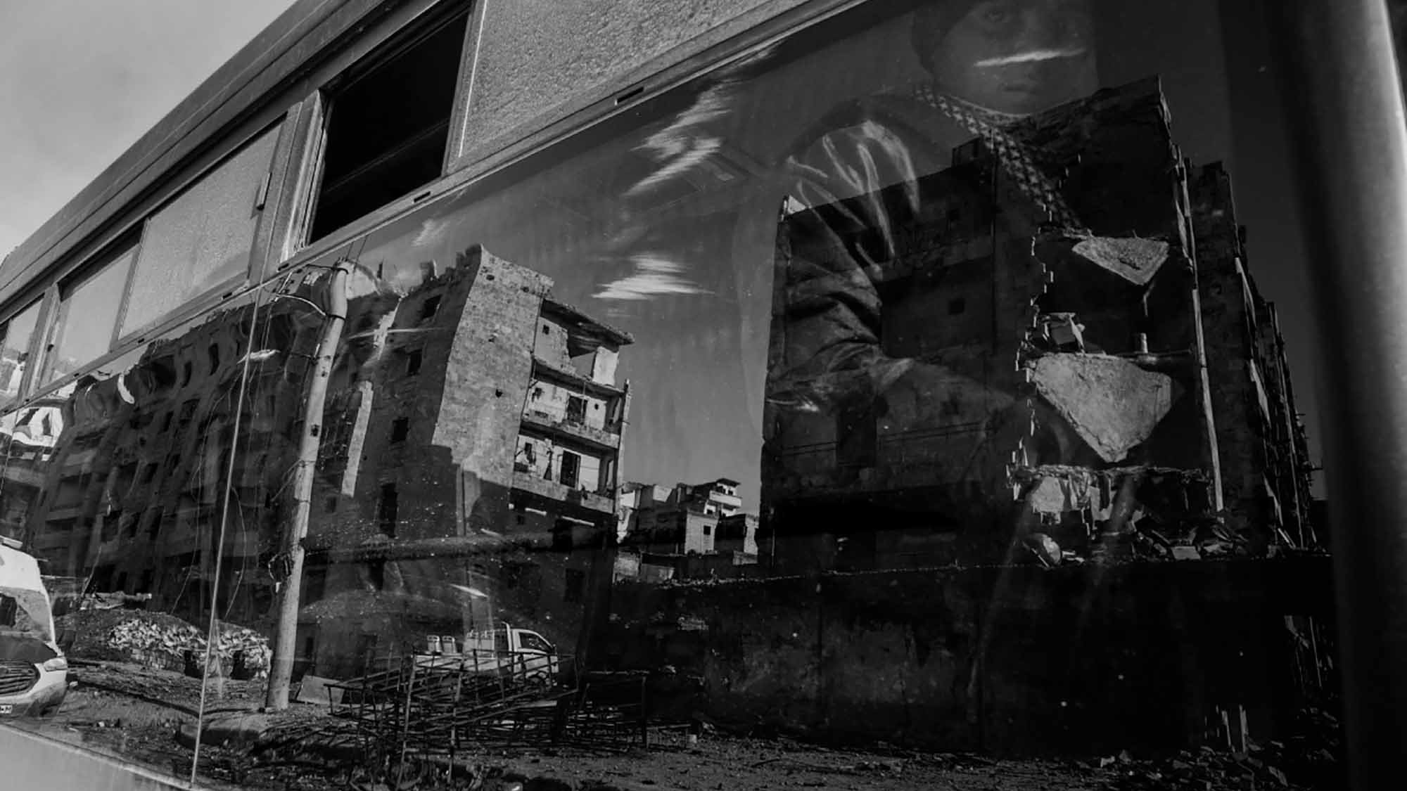 الصورة تظهر منظرًا حضريًا بواسطة زجاج نافذة حافلة. تعكس الصورة مجموعة من المباني المتضررة، مما يدل على دمار أو تدهور في المنطقة. يظهر أيضاً شخص يجلس بجانب النافذة، وملامح وجهه غير واضحة بسبب الانعكاس. الألوان في الصورة تتسم باللونين الأبيض والأسود، مما يضفي طابعاً درامياً وأجواءً حزينة على المشهد.