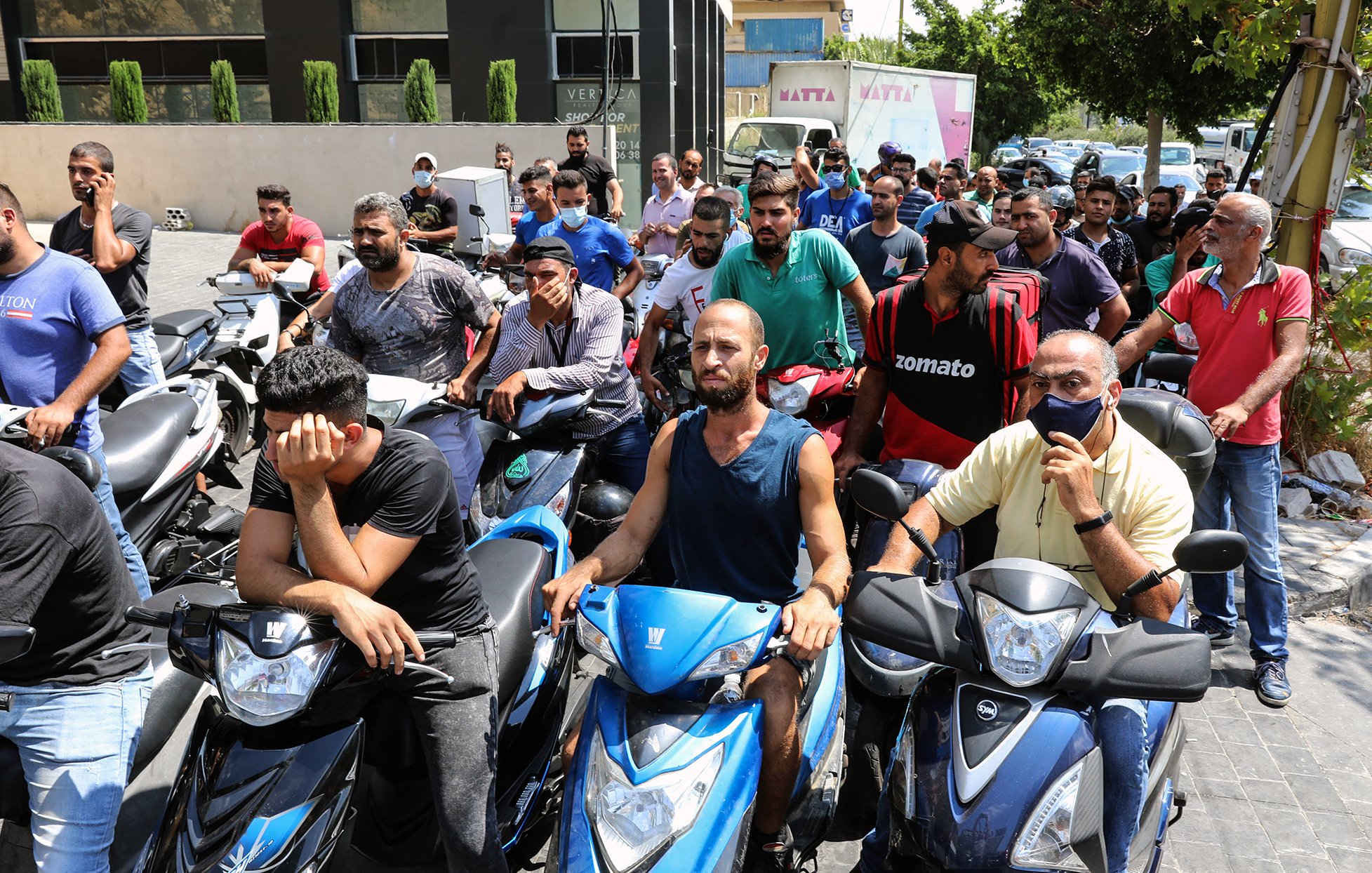 تظهر الصورة مجموعة من الأشخاص يتجمعون أمام مركز ما، حيث يبدو أنهم في انتظار الحصول على شيء ما. معظمهم يركبون دراجات نارية، ويبدو عليهم التوتر والقلق. بعضهم يظهر علامات الإحباط، بينما آخرون يتحدثون مع بعضهم. الخلفية تحتوي على مباني وأشجار، مما يشير إلى أن المشهد في منطقة حضرية.