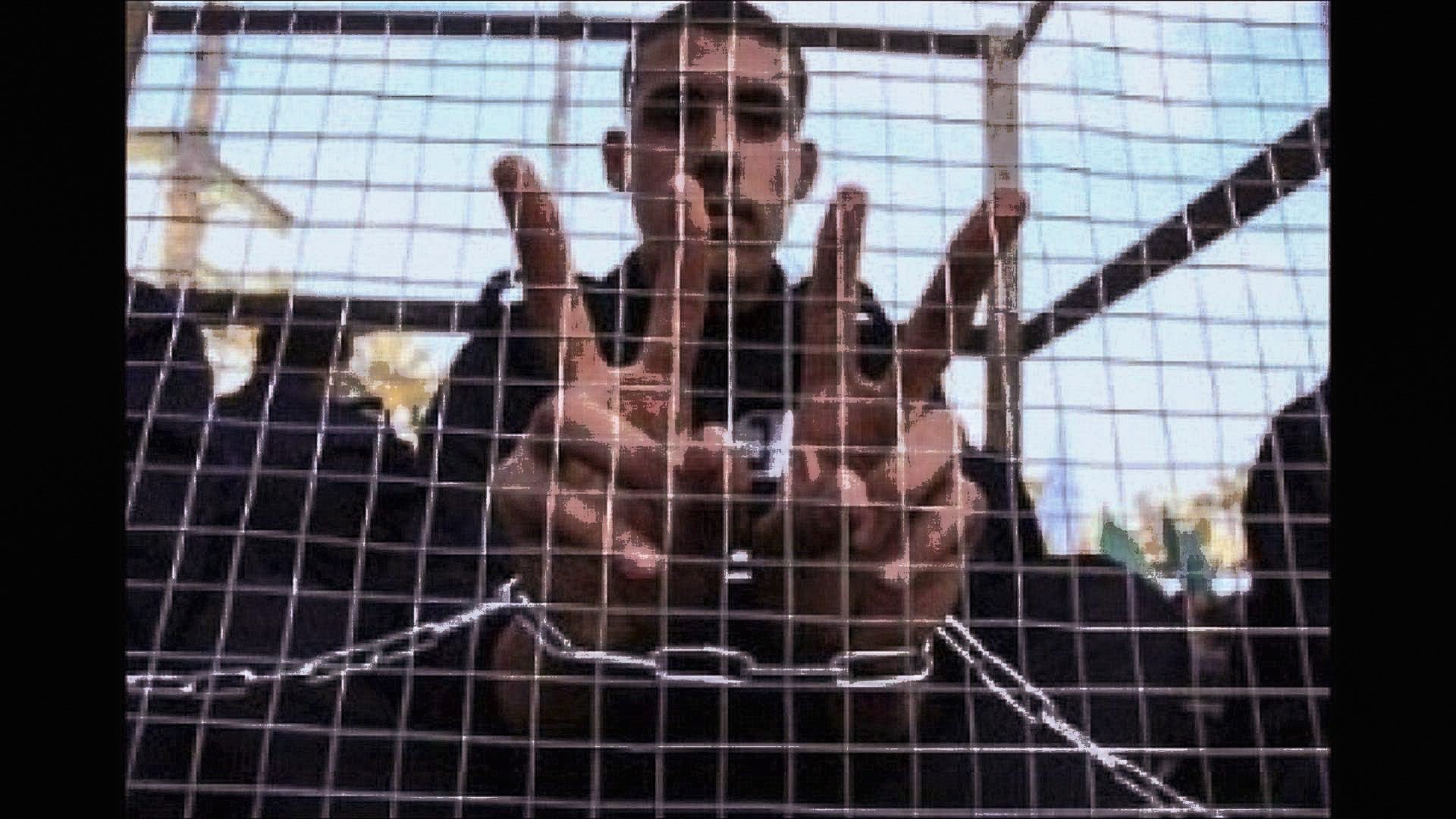 الصورة تظهر شابًا يقف خلف شبكة أو قفص، حيث يرفع يديه مكونًا علامة أو عددًا معينًا بإشارات أصابعه. يبدو أنه محاط بمساحة ضيقة، مما يعطي إيحاءً بالاحتجاز أو العزلة. تعكس تعابير وجهه أو وضع يديه مشاعر معينة، مثل المقاومة أو الإصرار. الخلفية غير واضحة تمامًا، مما يركز الانتباه على الشاب وإشارته.