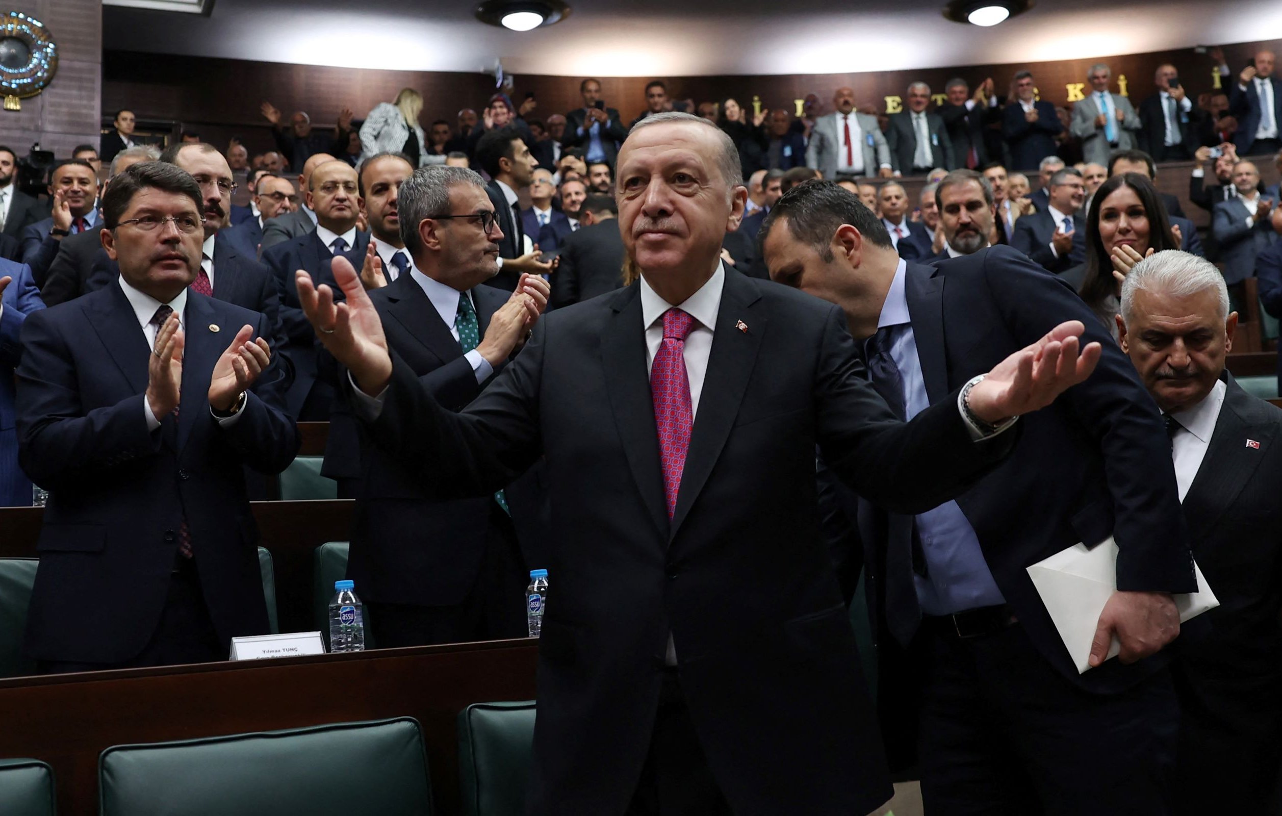الصورة تظهر رئيس تركيا، رجب طيب أردوغان، standing في مجلس يتكون من عدد كبير من الشخصيات. يبدو أنه يتحدث أو يقدم خطابًا، حيث يرفع ذراعيه بشكل يعبر عن التواصل أو الاستجابة. خلفه، يوجد مجموعة من الحضور الذين يصفقون أو يستمعون له، مما يوحي بأجواء من التفاعل والحماس. ترتدي الشخصيات خلفه ملابس رسمية، ويبدو أنهم في جلسة برلمانية أو حدث سياسي.