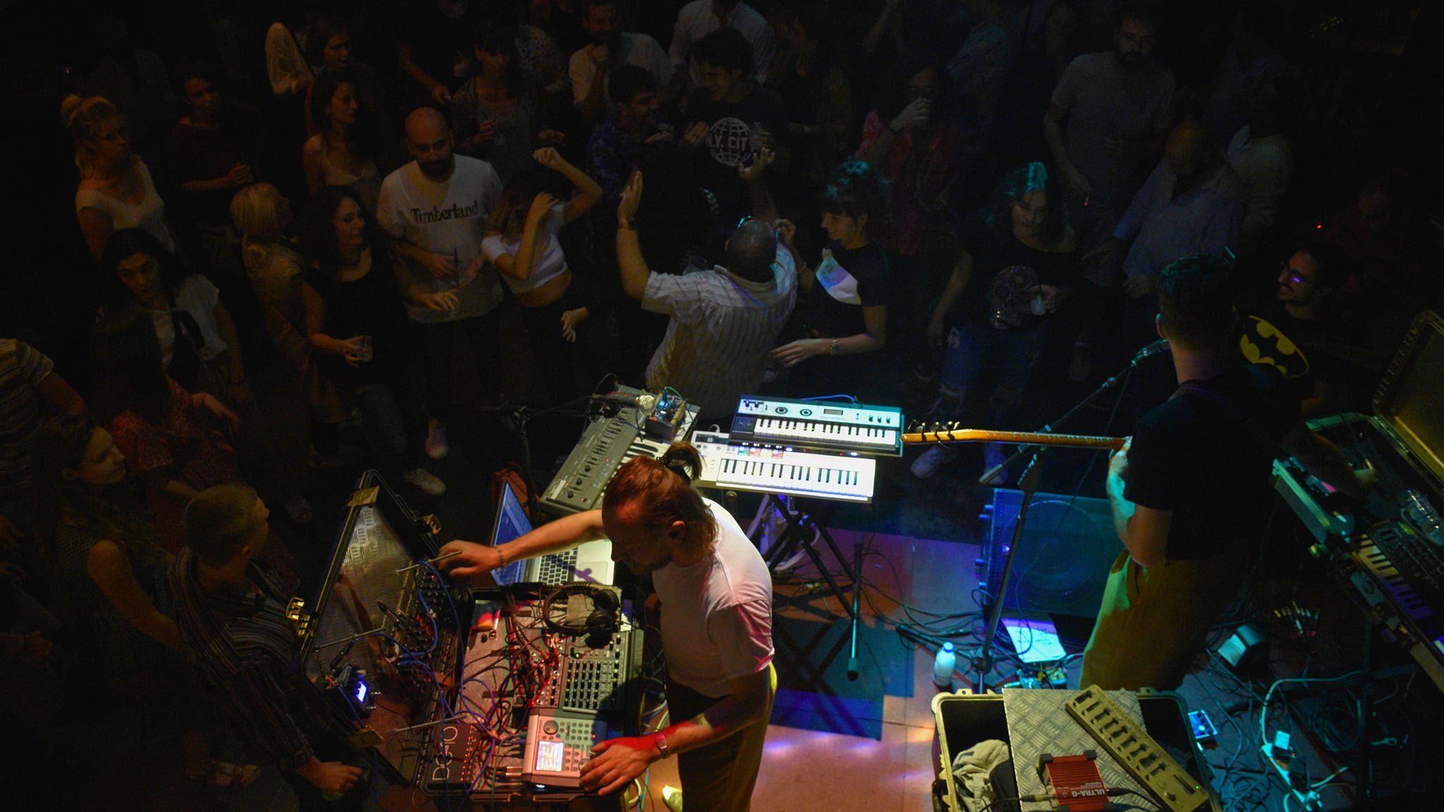 A lively crowd enjoys live music from performers with electronic instruments on stage.