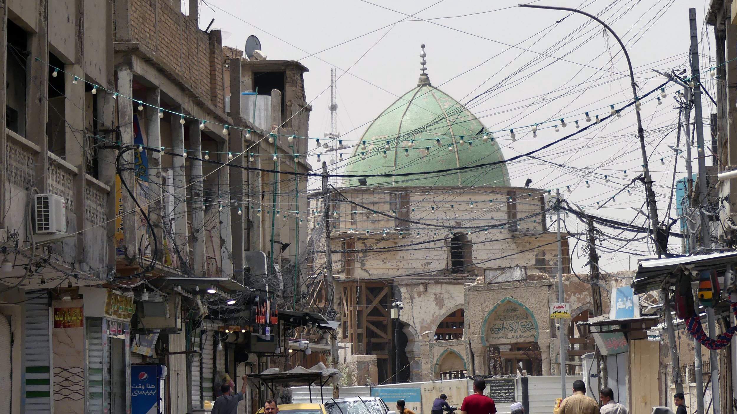 الصورة تظهر شارعًا في منطقة حضرية، حيث يتواجد عدد من الأشخاص يسيرون في اتجاهات مختلفة. في الخلفية، يظهر مسجد ذو قبة خضراء، مما يعكس الطابع المعماري المحلي. الشارع مليء بوسائل النقل، حيث توجد دراجة نارية وسيارة صفراء. بالإضافة إلى ذلك، تبرز الأسلاك الكهربائية المعلقة في الهواء والمحال التجارية على جوانب الطريق، مما يضفي على المشهد طابعًا حيويًا ونابضًا بالحياة.