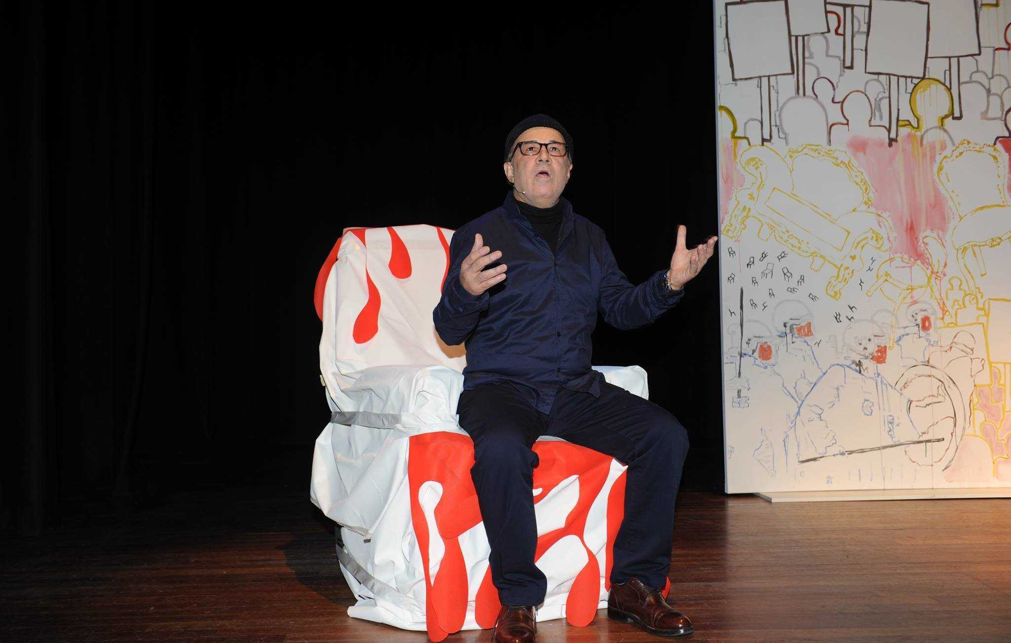 تظهر في الصورة شخصية جالسة على كرسي مغطى بقماش أبيض مزين بلمسات حمراء. يبدو أنه في وضع أداء أو حديث، حيث يعبّر باستخدام يديه ويظهر تركيزاً في تعبيراته. الخلفية تحتوي على لوحات جدارية غير محددة، مما يضفي جواً فنياً على المشهد. الشخص يرتدي ملابس داكنة ونظارات، ويبدو بشكل عام في حالة تفاعل مع الجمهور أو المشهد المحيط به.