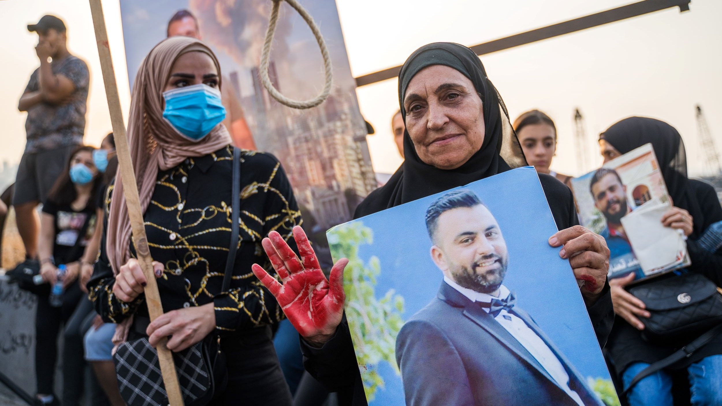 تظهر الصورة مجموعة من الأشخاص في مظاهرة. في المقدمة، هناك سيدة مسنّة ترتدي حجابًا وتمسك بصورة لرجل يبدو أنه قد توفي، بينما تضع يدها الملطخة باللون الأحمر، مما يوحي بمحتوى رمزي. بجانبها، توجد سيدة شابة ترتدي قناع وجه وأمسكت بلافتة. خلفهم، يظهر عدد من الأشخاص الذين يتفاعلون مع الحدث. وتبدو الأجواء مشحونة بالعواطف.