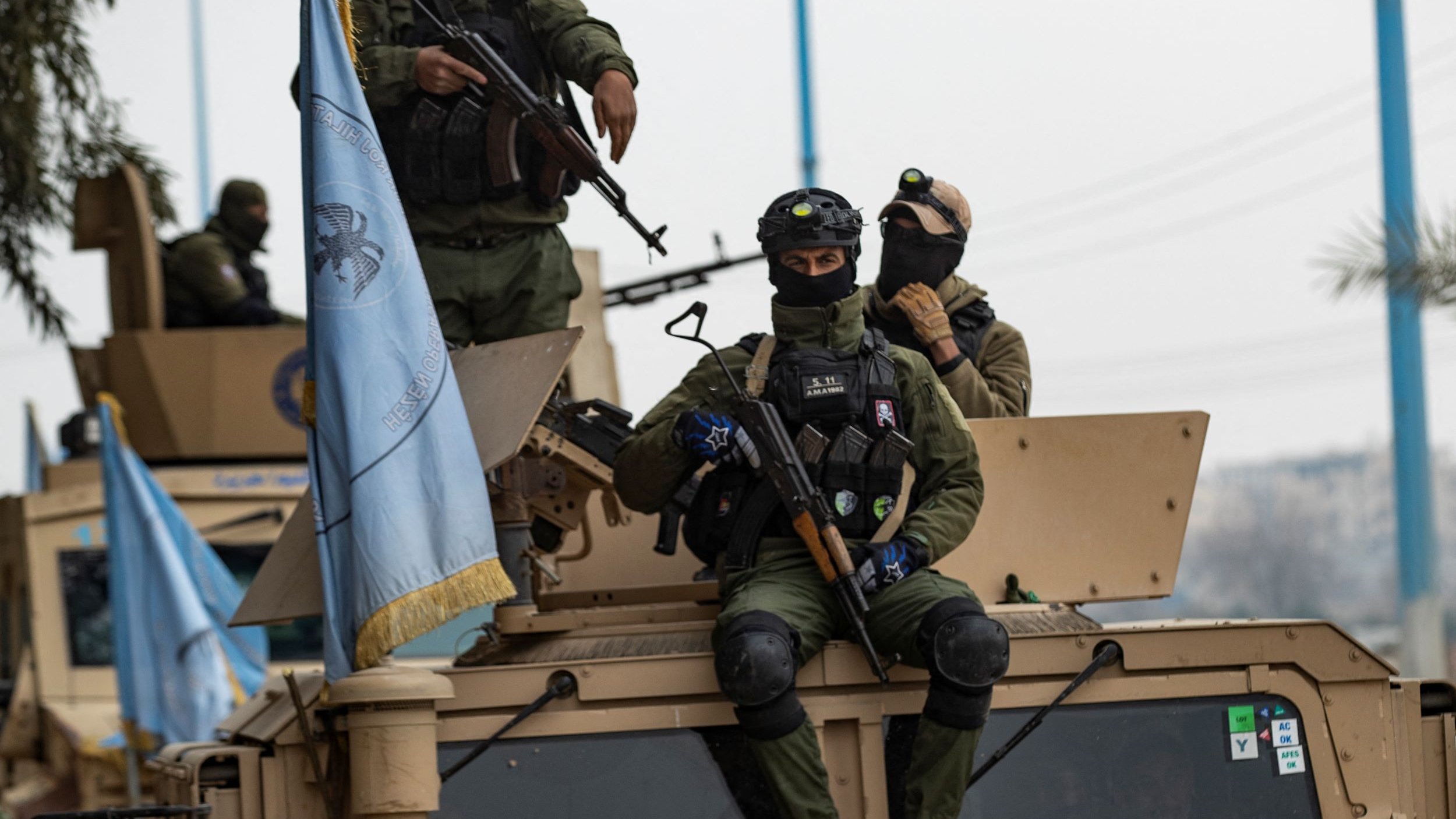 تظهر في الصورة مجموعة من الجنود العسكريين أثناء وجودهم في مركبة مدرعة. يرتدي الجنود زيًا عسكريًا ويظهر على بعضهم معدات تكتيكية وأسلحة. أحد الجنود يجلس على الجزء العلوي من المركبة، بينما يظهر آخرون يقفون عليها. هناك علم أزرق يعود لجهة معينة يبدو أنه متعلق بمهام حفظ السلام. الأجواء المحيطة تبدو حضرية مع وجود أشجار ومرافق عامة في الخلفية.