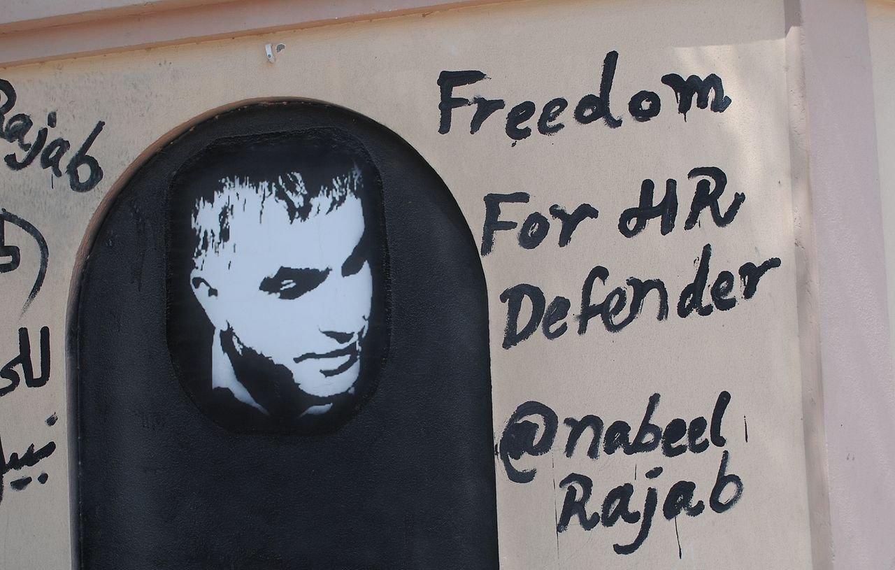الصورة تظهر جدارية على حائط، تحتوي على صورة لشخص يبدو وكأنه إنسان. تحت الصورة، هناك نص مكتوب باللغة الإنجليزية يقول "حرية لمدافع حقوق الإنسان" مع ذكر اسم "@nabeel Rajab". الألوان المستخدمة تتضمن الأسود والأبيض، حيث تبرز الصورة بشكل يلفت الانتباه. تعبر الجدارية عن دعم لشخصية معروفة في مجال حقوق الإنسان.