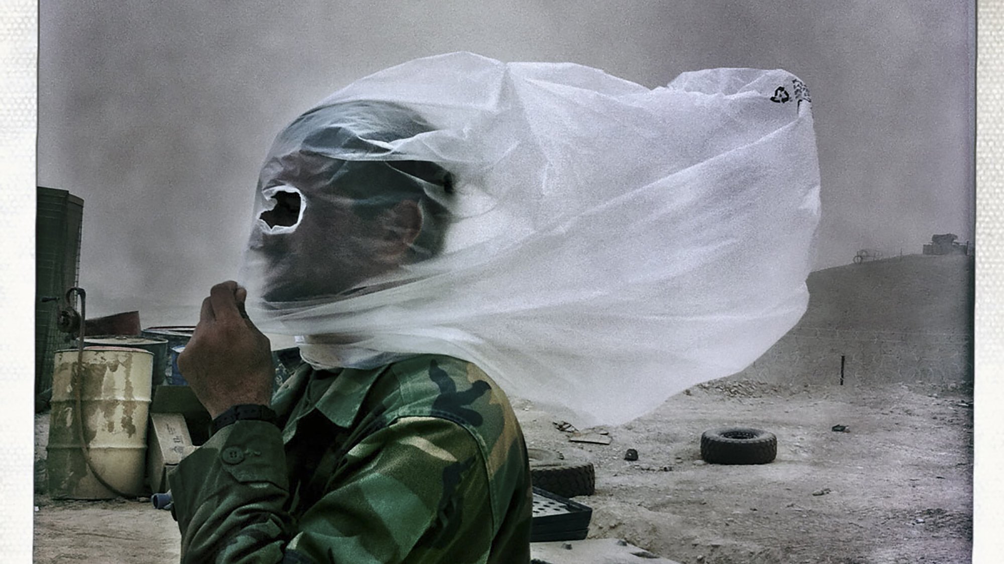 تظهر في الصورة شخص يرتدي زيًا عسكريًا، وقد غطى وجهه بقطع من القماش أو البلاستيك بسبب الظروف الجوية السيئة، مثل العواصف الرملية أو الغبار. يبدو أن الشخص واقف في منطقة غير مستوية، حيث يمكن رؤية بعض الأشياء في الخلفية مثل الإطارات والمواد الأخرى. تعكس الصورة حالة تحدٍ تحملها الشخص في مواجهة البيئة القاسية.