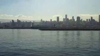 تظهر الصورة منظراً ساحلياً لمدينة حديثة، حيث يمتد الأفق في الخلفية مع مباني شاهقة. السطح المائي هادئ ويعكس ضوء الشمس، مما يخلق جوًا هادئًا. يبدو أن هناك سفينة راسية بالقرب من الميناء، بينما تظهر السحب الخفيفة في السماء فوق المباني. الألوان تبدو دافئة مع ظهور ضباب خفيف يعطي شعورًا بمسافة وتأمل في مشهد المدينة.