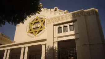 تظهر الصورة واجهة مبنى يمكن أن يكون معبدًا أو مركزًا ثقافيًا، يتميز بتصميم معماري فريد. الجدران الخارجية هي بألوان فاتحة، وهناك نجمة داود كبيرة مضاءة على الواجهة. يحيط بالمبنى أشجار خضراء، مما يضيف لمسة طبيعية. يبدو أن هناك نوافذ ذات تصميم زخرفي، مما يعطي المكان طابعًا مميزًا.