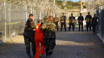 تظهر الصورة مشهدًا من مكان ذي طابع عسكري أو سجني. في المقدمة، هناك شخص يرتدي زي سجن برتقالي، ويبدو أنه يُقاد من قِبل اثنين من الجنود الذين يرتدون زيًا مموهًا. في الخلفية، يقف عدد من الجنود المرابطين، ويبدو أنهم في حالة تأهب. يحيط بالمكان سلك شائك، مما يدل على أنه منطقة محصورة أو محاطة بحراسة مشددة. السماء تبدو مشمسة، والأرض مغطاة بحصى.