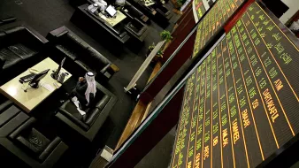 تظهر الصورة مشهداً من داخل سوق مالية أو بورصة، حيث يمكن رؤية شاشات إلكترونية كبيرة تعرض أسعار الأسهم ومعلومات السوق. في المقدمة، يوجد شخص يرتدي ملابس تقليدية يجلس أمام شاشة كمبيوتر، محاطاً بأثاث مريح يتضمن أرائك وطاولات. الأضواء والبيئة تعكس جو العمل والحركة في السوق المالية.