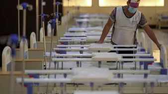 ترتدي الصورة مشهداً داخل مرفق صحي، حيث يظهر شخص واحد يرتدي كمامة وقميصاً. الشخص يركز على سرير طبي بين عدة أسرّة مرتبة بشكل منظم في غرفة واسعة مضاءة بشكل جيد. تظهر في الخلفية أعمدة ومعدات طبية، مما يوحي بأن المكان يُستخدم لعلاج المرضى. يتميز الجو بالهدوء والترتيب.