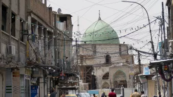 الصورة تظهر شارعًا في منطقة حضرية، حيث يتواجد عدد من الأشخاص يسيرون في اتجاهات مختلفة. في الخلفية، يظهر مسجد ذو قبة خضراء، مما يعكس الطابع المعماري المحلي. الشارع مليء بوسائل النقل، حيث توجد دراجة نارية وسيارة صفراء. بالإضافة إلى ذلك، تبرز الأسلاك الكهربائية المعلقة في الهواء والمحال التجارية على جوانب الطريق، مما يضفي على المشهد طابعًا حيويًا ونابضًا بالحياة.