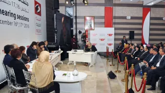 الصورة تظهر جلسة استماع تتعلق بموضوع اجتماعي أو سياسي. في المكان، يجلس عدد من الأشخاص على الجانبين، حيث يبدو أن هناك مجموعة من المتحدثين يجلسون على منصة مرتفعة. يظهر أمامهم طاولة مستديرة حيث يتم تقديم المداخلات. تُظهر الخلفية شعارات وصورة لمتحدث ما، مما يشير إلى أهمية الحدث. الأجواء تبدو رسمية ومرتبة، مع وجود حواجز حمراء تفصل بين الحضور.