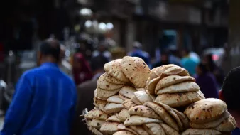 تظهر الصورة مجموعة من الخبز الطازج، يبدو أنه خبز عربي، مُتراص بشكل جميل في سلة يحملها شخص. في الخلفية، يتواجد عدد من الأشخاص في شارع مزدحم، مما يوحي بأجواء نابضة بالحياة في المدينة. يركز المشهد على الخبز كجزء من الثقافة الغذائية.