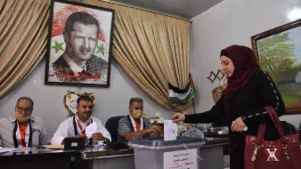 تظهر الصورة امرأة ترتدي عباءة وحجابًا تضع ورقة اقتراع في صندوق الانتخاب. خلفها، يوجد لوحة كبيرة تحمل صورة شخصية معروفة. يجلس ثلاثة رجال خلف طاولة، حيث يبدو عليهم الاستعداد لمراقبة العملية الانتخابية. المكان يبدو رسميًا، مع ستائر وخلفية تشير إلى الطابع المحلي.