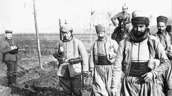 الصورة تظهر مجموعة من الجنود يسيرون في منطقة مفتوحة، يبدو أن الأرض مبللة ومليئة بالطين. الجنود يرتدون زيًا عسكريًا تقليديًا، بعضهم يحمل أسلحة، بينما يبدو أحدهم راكبًا على حصان. يمكن رؤية شخص آخر في الخلفية يقف على الجانب. الأجواء العامة توحي بأنها قد تكون خلال فترة تاريخية معينة من الحرب أو النزاع.