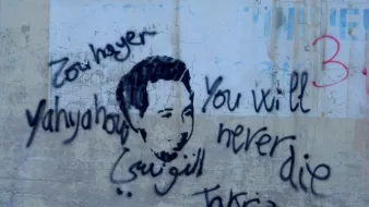 تظهر الصورة رسمًا على جدار يظهر وجه شخص، محاطًا بعبارات مكتوبة بخط يدوي. من بين هذه العبارات، تُظهر باللغة الإنجليزية "You will never die" (لن تموت أبدًا) وفي اللغة العربية "يحيى" و"تقديس". يبدو أن العمل يعبر عن رسالة تعاطف أو تأبين، مع تداخل بين الثقافات واللغات في الكتابة.