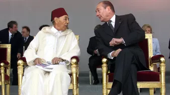 الصورة تظهر شخصين يجلسان على كراسي فاخرة. يبدو أن الحديث يجري بينهما بشكل ودي. الشخص الأول يرتدي زيًا تقليديًا مغربيًا مع طربوش، بينما الشخص الثاني يرتدي بدلة رسمية داكنة. في الخلفية توجد مجموعة من الأشخاص الجالسين، ويبدو أنهم في مناسبة رسمية أو احتفال. الضوء في الصورة ناعم، مما يضيف جوًا من الألفة.