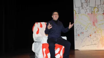 تظهر في الصورة شخصية جالسة على كرسي مغطى بقماش أبيض مزين بلمسات حمراء. يبدو أنه في وضع أداء أو حديث، حيث يعبّر باستخدام يديه ويظهر تركيزاً في تعبيراته. الخلفية تحتوي على لوحات جدارية غير محددة، مما يضفي جواً فنياً على المشهد. الشخص يرتدي ملابس داكنة ونظارات، ويبدو بشكل عام في حالة تفاعل مع الجمهور أو المشهد المحيط به.