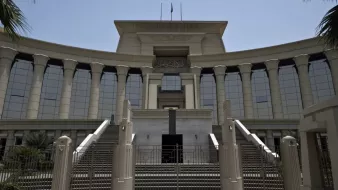 الصورة تظهر مبنى حكومي كبير يتميز بتصميم معماري حديث. يبرز في الصورة مدخل المبنى الذي يحتوي على مجموعة من السلالم والأعمدة الكبيرة. الواجهة تمتاز بالنوافذ الزجاجية العريضة التي تعكس الضوء، وتوجد أعلام مرفوعة فوق السطح. المنطقة المحيطة بالمبنى تحتوي على أشجار ونباتات تعطي جواً من الخضرة. يبدو الطقس مشمساً وواضحاً، مما يضفي على المشهد حيوية.