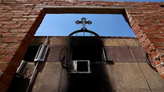 الصورة تظهر إطارًا من الطوب الأحمر مع فتحة في الجزء العلوي، ومن خلال هذه الفتحة يمكن رؤية صليب مرفوع في السماء. الجدران المحيطة تبدو متضررة، حيث يظهر عليها أثر السواد، مما يشير إلى وجود حريق أو تلف. السماء في الخلفية زرقاء وصافية، مما يعكس تناقضًا مع الأجواء المحيطة.