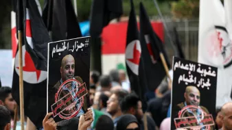 الصورة توضح تجمعًا حاشدًا حيث يحمل المتظاهرون لافتات تحمل عبارات وشعارات مختلفة. من بين اللافتات، توجد صور لشخص يبدو أنه شخصية بارزة، مع وجود علامة تشير إلى معارضة أو احتجاج يتعلق بموضوع الإعدام. المتظاهرون يرتدون ملابس متنوعة، وبعضهم يحمل أعلامًا. الأجواء تعكس شعوراً بالاحتجاج والرفض.