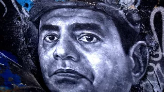 تظهر الصورة رسما جداريًا لوجه رجل، حيث يتميز التعبير بالعمق والتفاصيل الدقيقة. يُظهر الرسم عيونًا تعكس مشاعر قوية، والخطوط تتباين بين الظل والنور، مما يعطي إحساسًا بالحياة والواقعية. الخلفية تحتوي على ألوان داكنة ومخططات فنية أخرى، مما يبرز التركيز على وجه الرجل ويعزز من جمال العمل الفني.