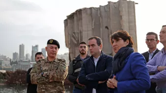 تظهر في الصورة مجموعة من الأشخاص في منطقة تضررت من انفجار أو حدث كارثي. في المقدمة، يوجد ضابط عسكري يرتدي زيًا رسميًا ولديه شارة على القبعة. بجانبه، يقف مجموعة من الرجال والنساء يتحدثون بجدية، مع تعابير وجه تعكس الانتباه والتفكير. في الخلفية، توجد آثار دمار واضحة، مع مبنى شاهق متضرر بشكل كبير. السماء تميل إلى اللون الرمادي، مما يضفي أجواءً من الحزن أو التأمل على المشهد.