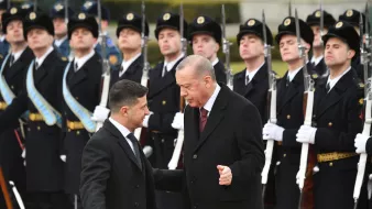 تظهر الصورة مجموعة من الجنود في الزي الرسمي يتواجدون في تشكيل عسكري أمام شخصيتين بارزتين. الشخصيات تبدو وكأنها تتحدثان معًا، حيث يبدو أحدهما في حالة نقاش مع الآخر. الجنود خلفهم يقفون بانضباط، بينما يتم عرض الأسلحة بشكل منظم. الأجواء تعكس طابعًا رسميًا وجديًا للحدث.