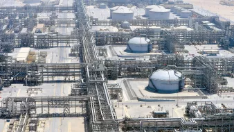 الصورة تُظهر منظرًا جويًا لمصنع صناعي ضخم، يبدو أنه يتعلق بقطاع النفط أو الغاز. يمكن رؤية مجموعة من الخزانات الدائرية الكبيرة، بالإضافة إلى شبكة معقدة من الأنابيب والبنى التحتية الأخرى. المنطقة محاطة بأرض صحراوية، مما يدل على أن المصنع يقع في منطقة جافة. يركز التصميم على الكفاءة في نقل وإدارة المواد الخام، مما يشير إلى مستوى عالٍ من التكنولوجيا والهندسة.