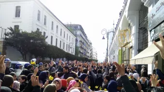 الصورة تظهر تجمعًا حاشدًا من الناس في مكان عام، حيث يرفع المشاركون أيديهم في الهواء. يبدو أن المتظاهرين يجسدون روح الاعتراض أو الدعوة إلى التغيير. الخلفية تحتوي على مبانٍ عالية، مما يعكس أجواء المدينة. يوجد أيضًا بعض رجال الشرطة في الصورة، مما يشير إلى أن الحدث قد يكون مظاهرة أو احتجاجًا. الأجواء تبدو مشحونة بالحماس والنشاط.